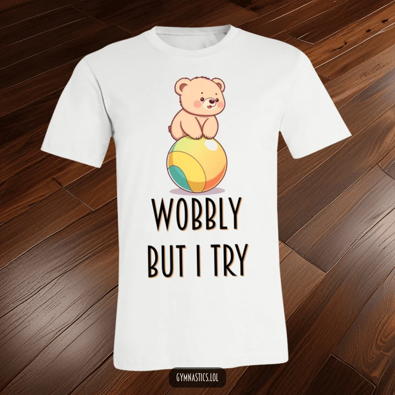 Funny t-shirt depicting a fluffy bear cub wobbling precariously on a colorful ball, showcasing adorable effort.